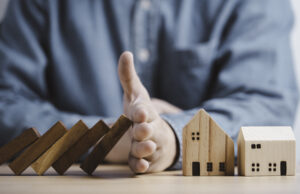 Property owner's hand protects and stops the wooden house from dominos falling on it for risk management.