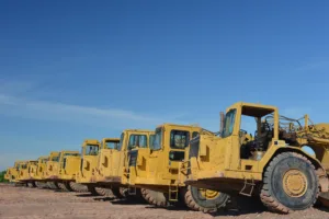 A row of yellow tractors securely stored on a job site during off-hours. They sit parked on the dirt, protected by security cameras, fencing, and access control that are located out-of-frame.