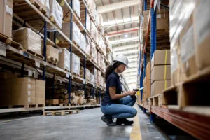 Woman kneeling down in a warehouse infront of some shelves of boxes taking inventory.